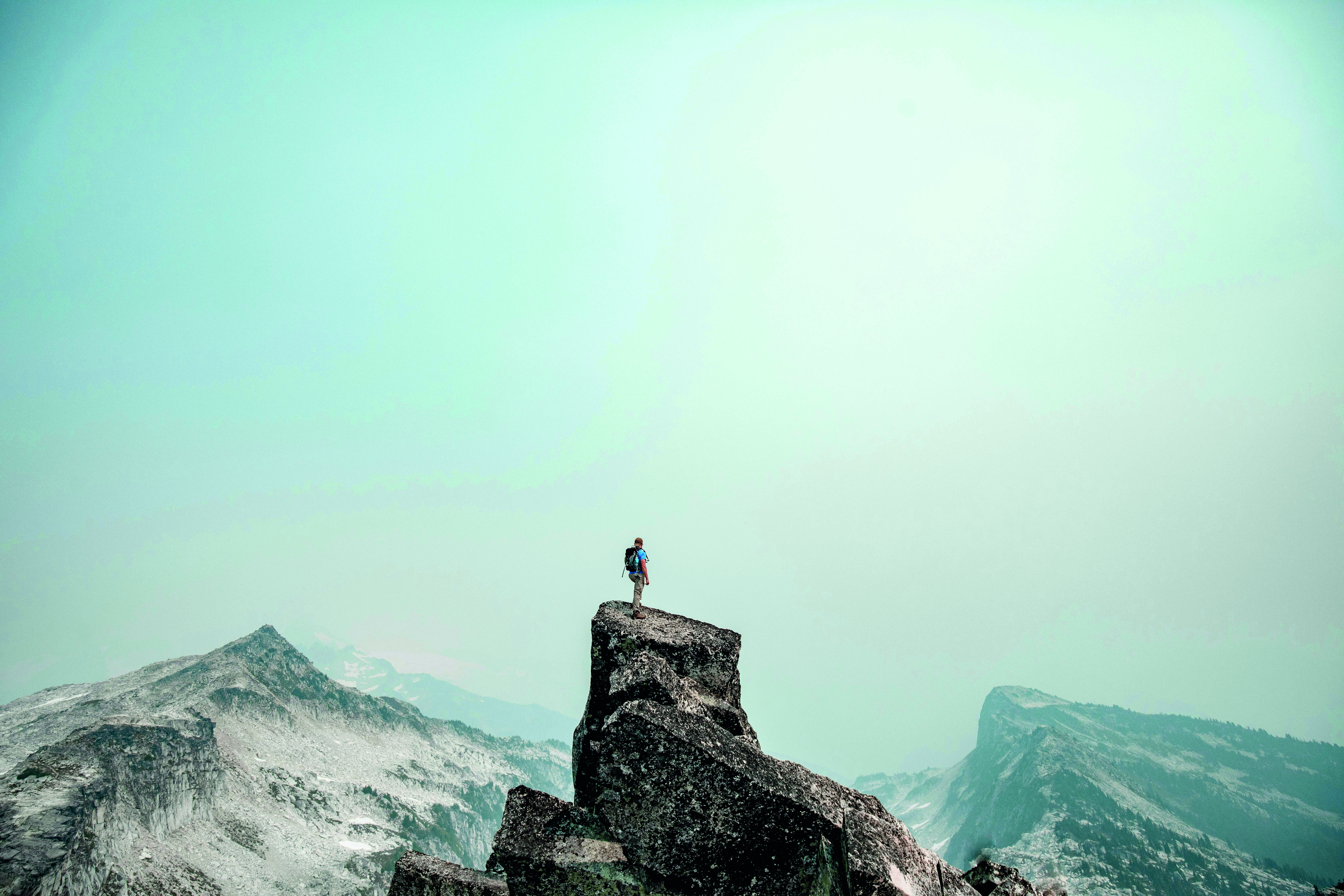 Hiker with backpack standing on a rocky mountain peak, surrounded by snow-patched mountains under a clear sky.