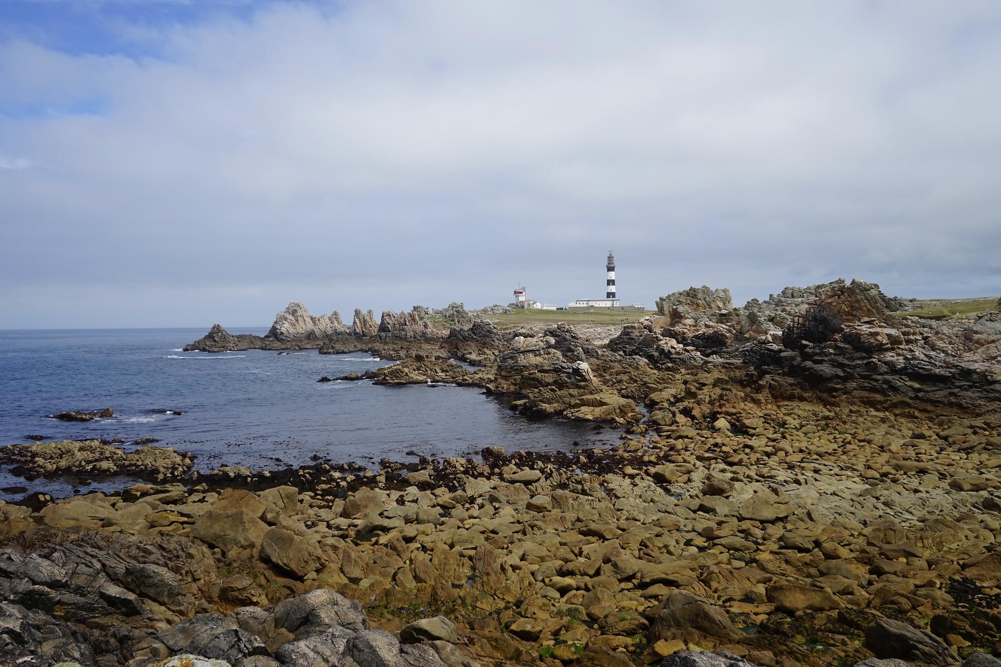 Landschaftsansicht der Insel Ouessant mit dem historischen Leuchtturm über der felsigen Küste und dem ruhigen Meer.