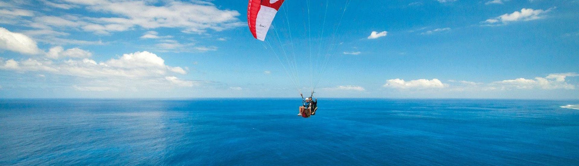 A paraglider pilot from Addict Parapente is doing a tandem paragliding flight in the blue sky of Reunion Islan