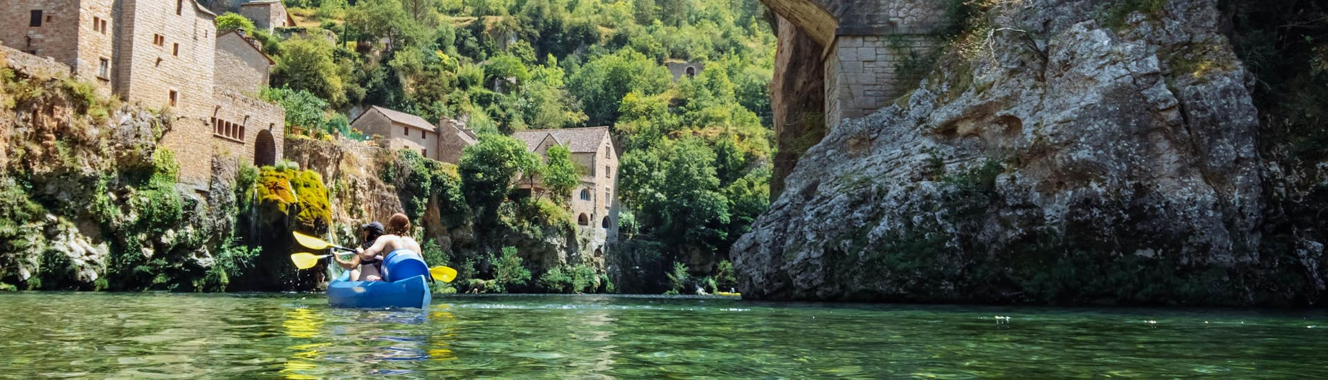 Two people are paddling down the Tarn River while crossing a bridge in the famous medieval town of Sainte-Énimie in the Tarn Gorges, one of France's popular canoeing locations.