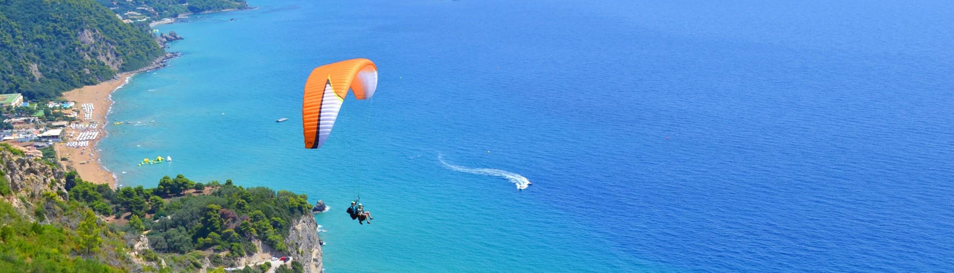 Ein  Tandempilot von Corfu Paragliding und sein Passagier gleiten bei einem Tandemflug über das türkisfarbene Wasser des Ionischen Meeres.