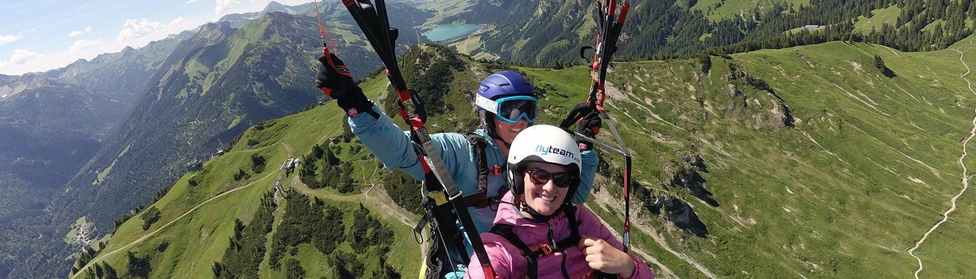 A tandem pilot from FlyTeam and her passenger are flying over the green mountain landscape during a tandem paragliding flight.
