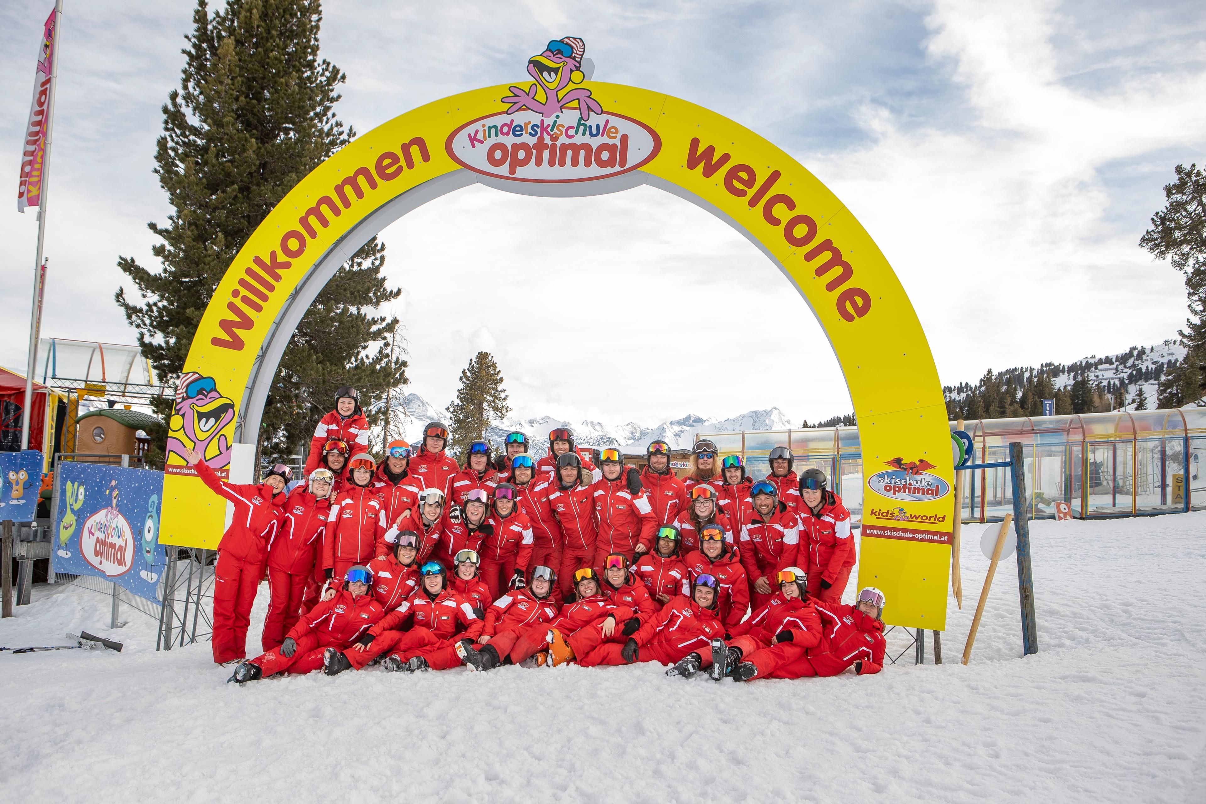 Een foto van alle skileraren van Skischule Optimal Kaltenbach voor een grote boog, met de bergen op de achtergrond.
