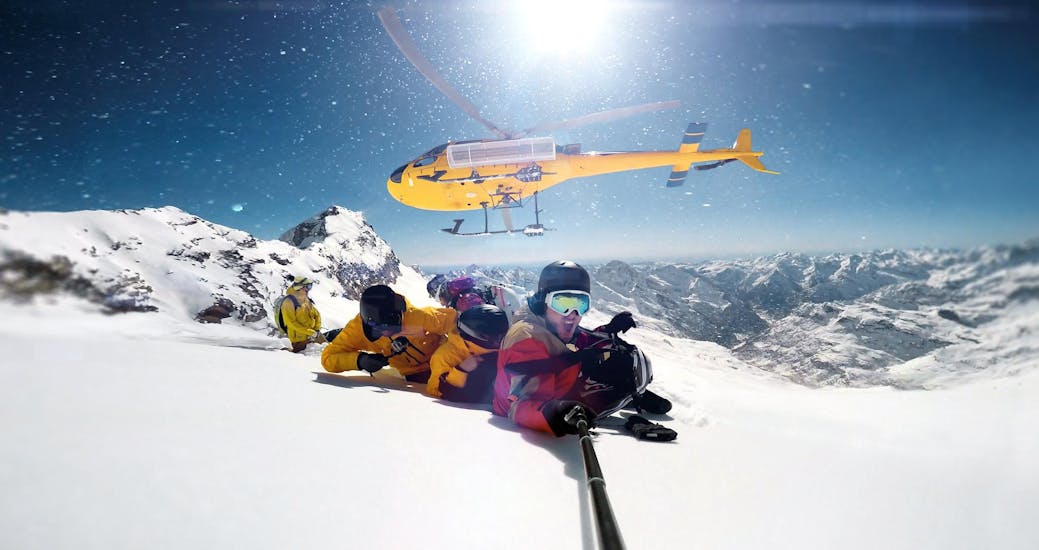 Eine Gruppe junger Menschen liegt bei der Aktivität "Heliskiing" mit dem Anbieter Christoph Zangerl - ifreeride im Schnee am Gipfel eines Berges und macht ein Selfie mit dem Helikopter im Hintergrund. 