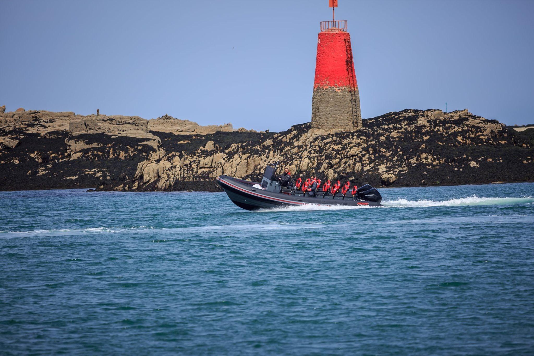 Le capitaine des Vagabonds de la Baie Roscoff sur son bateau lors d'une balade dans en mer avec au loin un phare et des falaises.