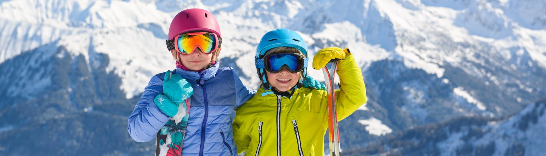 Two children in full skiing gear are smiling at the camera as they prepare for their kids ski lessons on top of the mountain.
