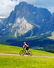Un uomo, mentre va in mountain bike sull'Alpe di Siusi, percorre un prato verde ai piedi di un'alta catena montuosa.
