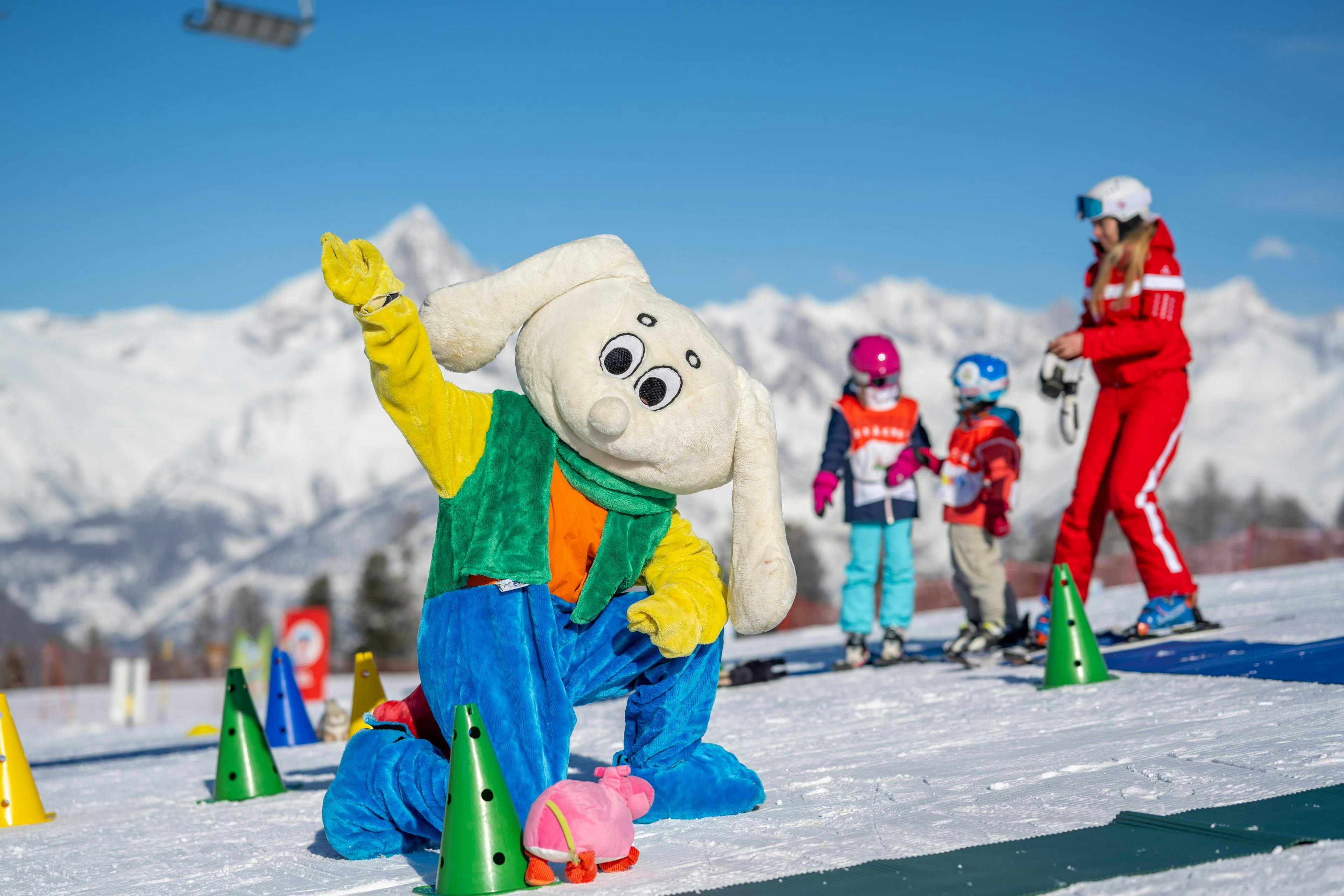 Swiss Ski and Snowboard School Grächen La mascotte "Snowli" e due piccoli sciatori con il loro maestro di sci durante una lezione di sci per bambini della Schweizer Ski- und Snowboardschule Grächen con uno sfondo montuoso.
