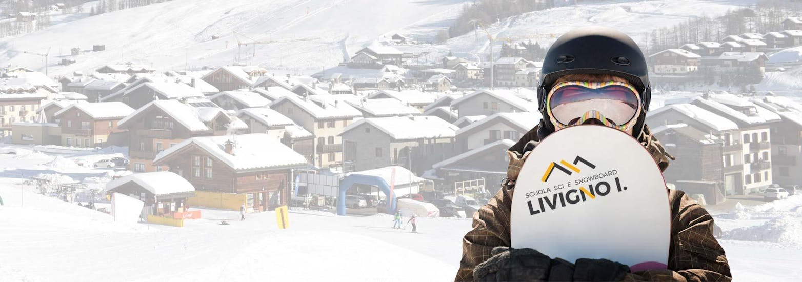 Ein Snowboarder der mit der Skischule Scuola di Sci e Snowboard Livigno Italy das Snowboarden lernt blickt in die Kamera mit der Ortschaft Livigno im Hintergrund.