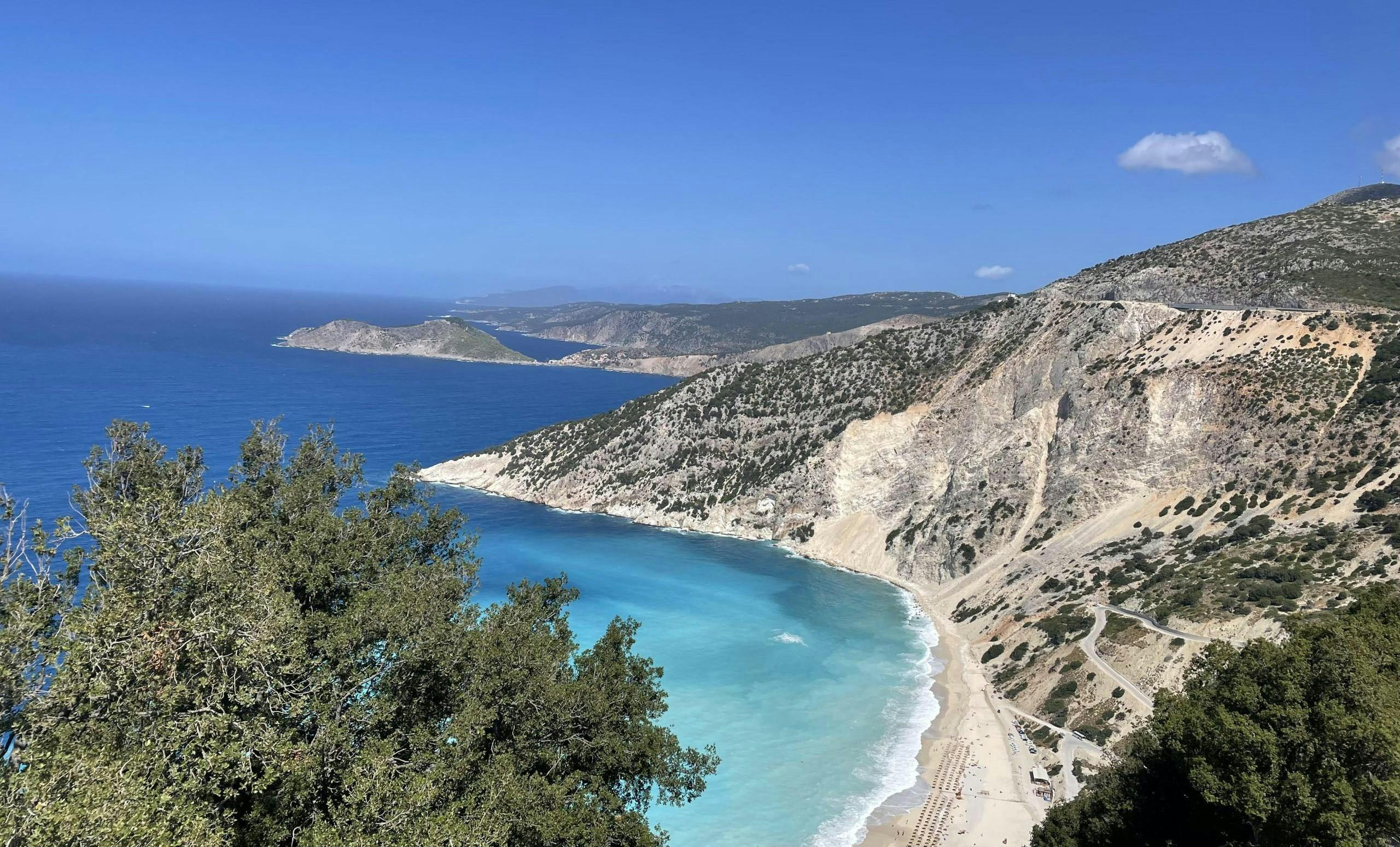 Sea Dream Travel Kefalonia Una vista costera pintoresca con una bahía turquesa, una playa de arena y acantilados escarpados bajo un cielo azul claro.