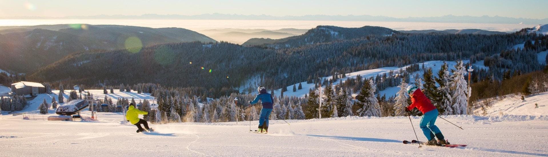 Zwei Skifahrer und ein Snowboarder ziehen auf der Skipiste in Feldberg im Schwarzwald, wo die örtlichen Skischulen ihre Skikurse anbieten, ihre Spuren.