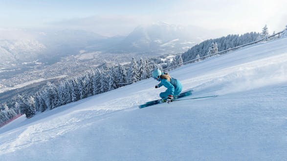 Eine Skifahrerin zieht eine perfekt präparierte Piste mit Blick auf das Skigebiet Garmisch-Classic hinunter, wo auch die örtlichen Skischulen eine Bandbreite an Skikursen anbieten. (c)Bayerische Zugspitzbahn Bergbahn AGfendstudios.com