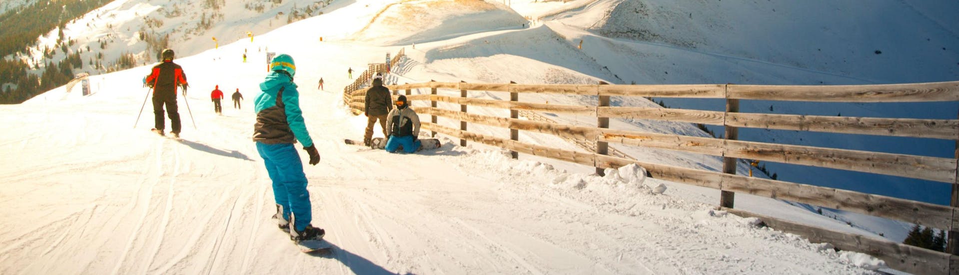 Eine Gruppe Skifahrer und Snowboarder fährt eine in Sonnenlicht getränkte Skipiste im Skigebiet Hinterglemm hinunter, wo örtliche Skischulen ihre Skikurse für die die das Skifahren lernen wollen anbieten.