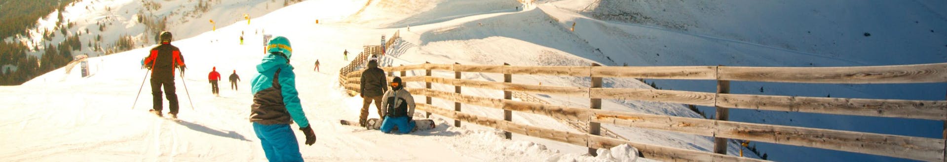 Eine Gruppe Skifahrer und Snowboarder fährt eine in Sonnenlicht getränkte Skipiste im Skigebiet Hinterglemm hinunter, wo örtliche Skischulen ihre Skikurse für die die das Skifahren lernen wollen anbieten.