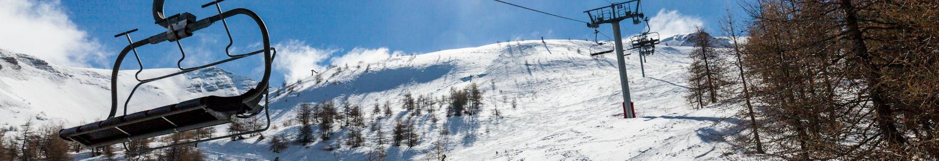 Ein Bild eines Sessellifts im Skigebiet von Les Orres, neben dem eine Gruppe Skifahrer die an einem von einer örtlichen Skischule organisierten Skikurs teilnimmt die Piste hinab fährt.