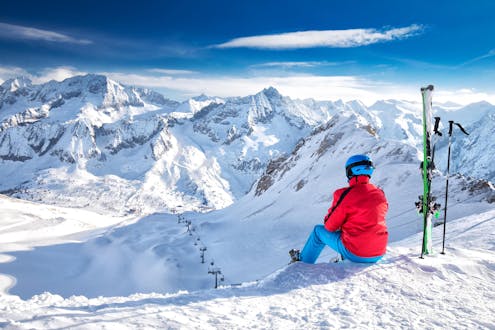 Ein Skifahrer sitzt oben auf der Skipiste im Schnee und blickt hinunter auf das Skigebiet Passo del Tonale, wo Besucher bei einer der örtlichen Skischulen einen Skikurs buchen können.