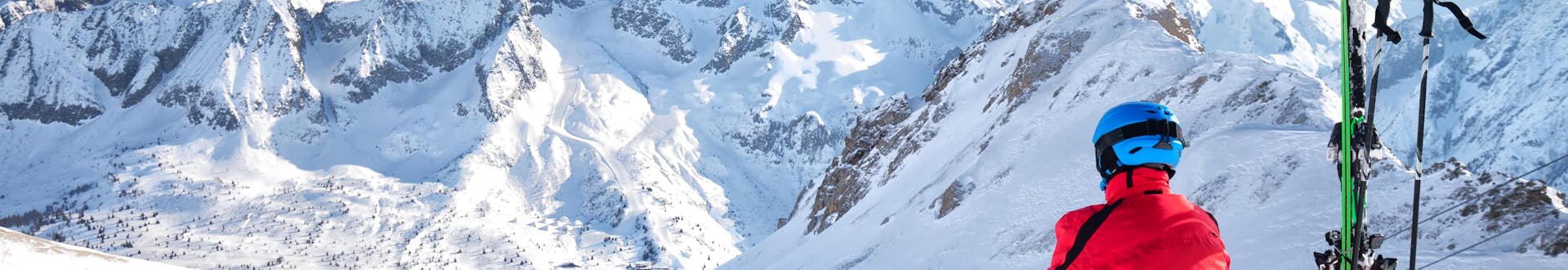 A skier is sitting in the snow at the top of the ski slope, looking down over the ski resort of Passo del Tonale, where visitors can book ski lessons with one of the local ski schools.