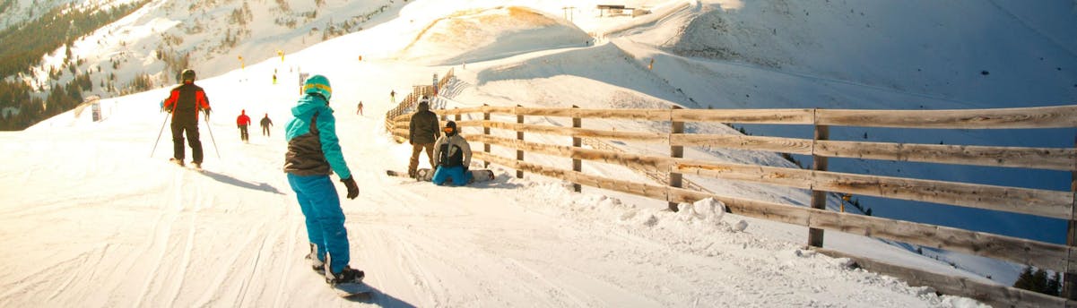 ski-school-saalbach_SEM-Resort-Hero A couple of skiers and snowboarders are riding down a sunlit skislope in the ski resort of Saalbach, where local ski schools offer ski lessons for people who want to learn to ski.