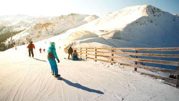 Eine Gruppe Skifahrer und Snowboarder fährt eine in Sonnenlicht getränkte Skipiste im Skigebiet Saalbach hinunter, wo örtliche Skischulen ihre Skikurse für die die das Skifahren lernen wollen anbieten.