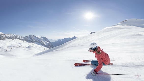 A female skier is skiing in front of a stunning backdrop of snowy mountain tops glistening in the sunshine in the ski resort of Zugspitze, where the local ski schools offer their ski lessons. (c)Bayerische Zugspitzbahn Bergbahn AGfendstudios