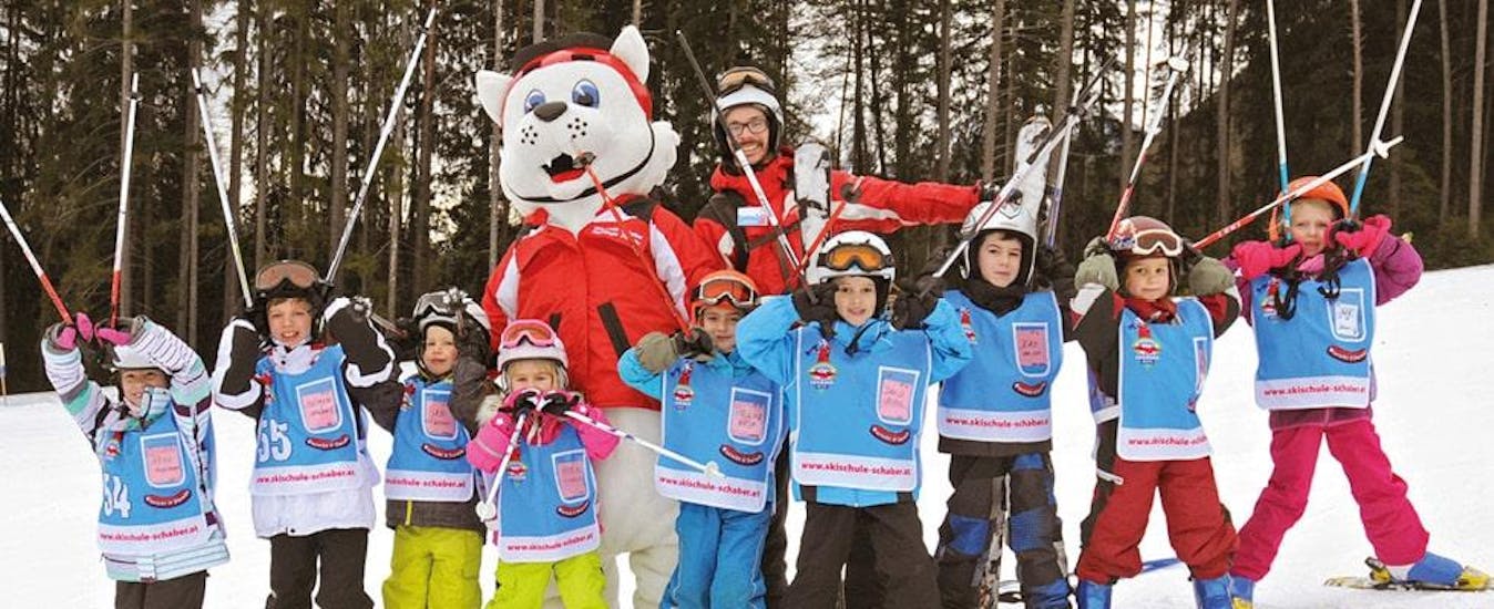 Eine Gruppe junger Kinder posiert bei ihrem Kinder-Skikurs gemeinsam mit ihrem Skilehrer der Skischule Schaber in Grünberg Obsteig und dem Skischul-Maskottchen Hermi Hermelinchen für ein Foto.