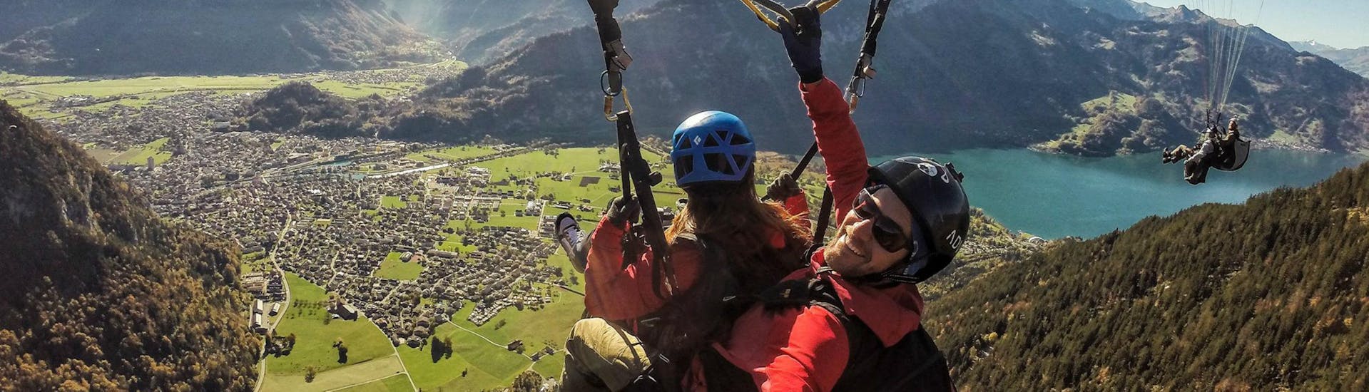 A tandem pilot from Skywings Interlaken is taking a selfie while his passenger enjoys the panoramic view of the Swiss mountains during their paragliding flight.
