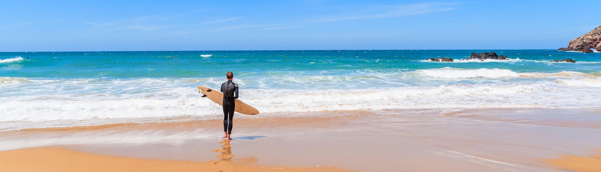 Ein junger Mann in einem Neoprenanzug steht beim Surfen in Arrifana mit seinem Surfbrett an einem Strand und blickt auf den Ozean.