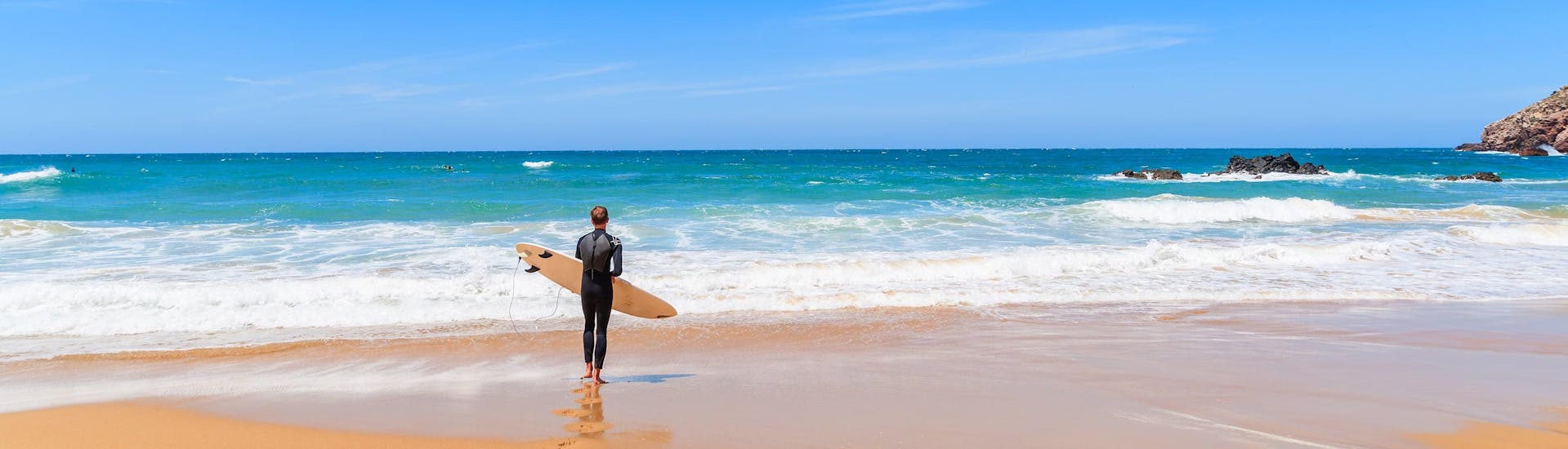 Ein junger Mann in einem Neoprenanzug steht beim Surfen in Aljezur mit seinem Surfbrett an einem Strand und blickt auf den Ozean.