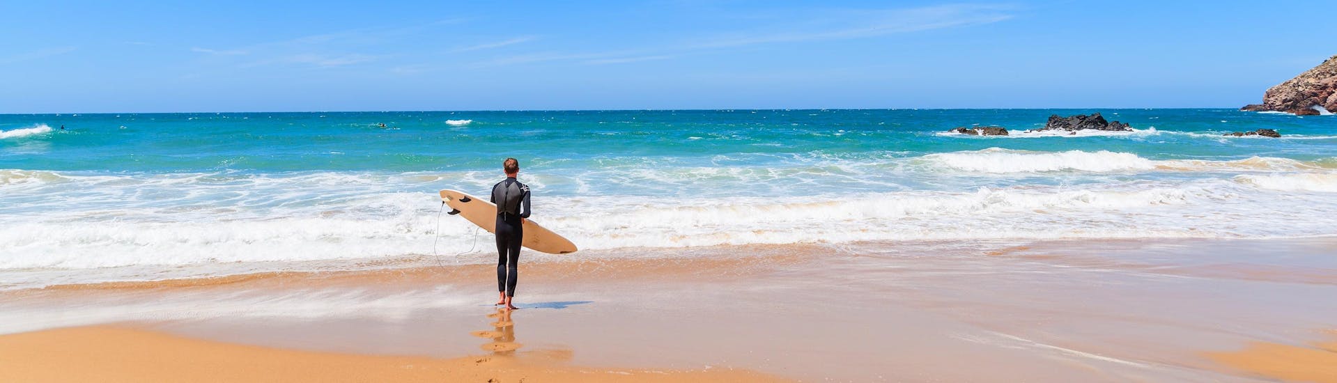 A young man dressed in a wetsuit is standing on a beach with his surfboard, looking out over the ocean as he gets ready to go surfing in the Algarve.