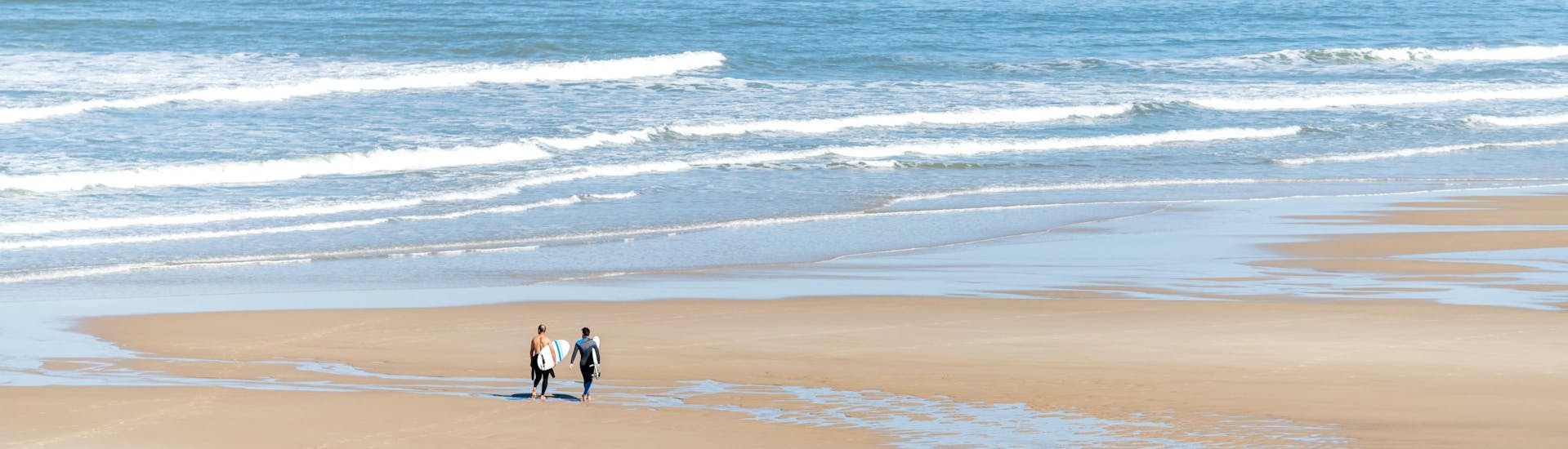 Deux hommes marchent sur la plage centrale de Lacanau avec leur planche de surf sous le bras, où se déroulent de nombreux cours de surf.