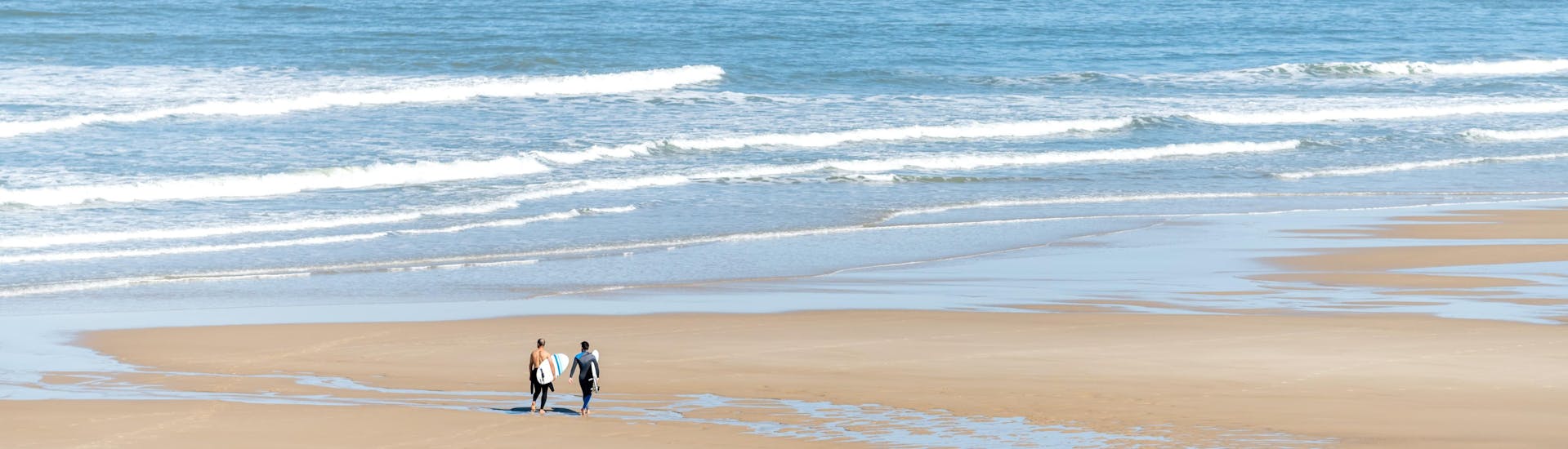 Deux hommes marchent sur la plage centrale de Lacanau avec leur planche de surf sous le bras, où se déroulent de nombreux cours de surf.