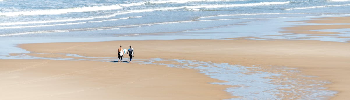 surfing-lacanau_SEM-Resort-Hero Two men are walking on the beach of Lacanau with their surfboard under their arm, where many surfing lessons take place.
