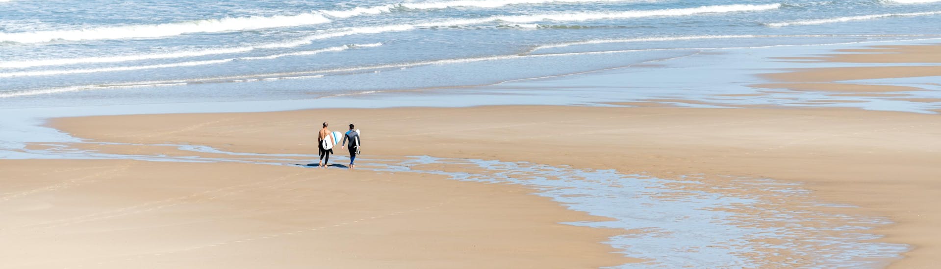 Two men are walking on the beach of Moliets-et-Maâ with their surfboard under their arm, where many surfing lessons take place.