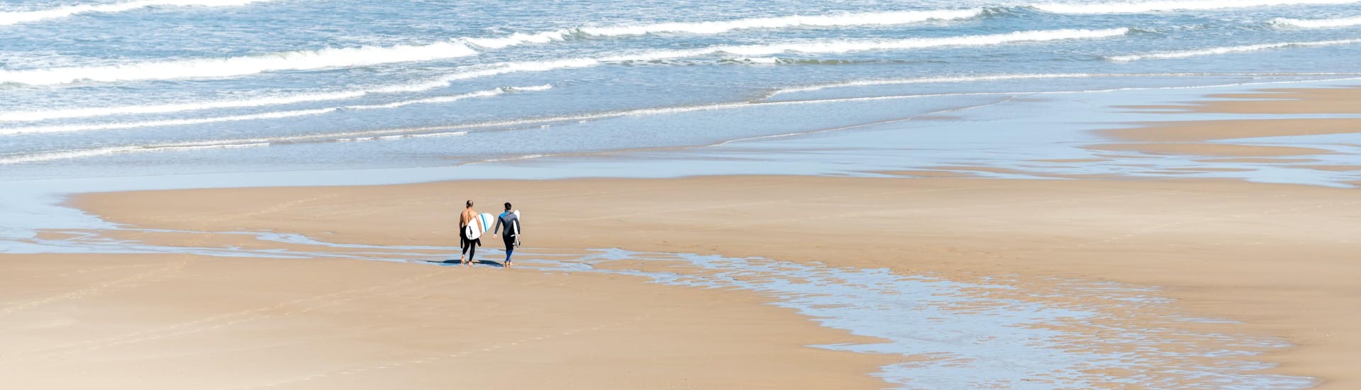 Deux hommes marchent sur la plage de Lacanau avec leur planche de surf sous le bras, où se déroulent de nombreux cours de surf.