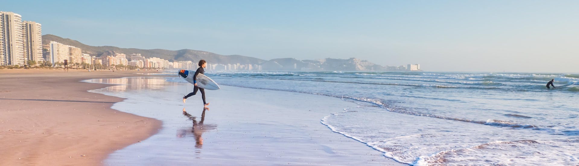 Un hombre joven corre hacía el oceáno mientras hace surf en Valencia.