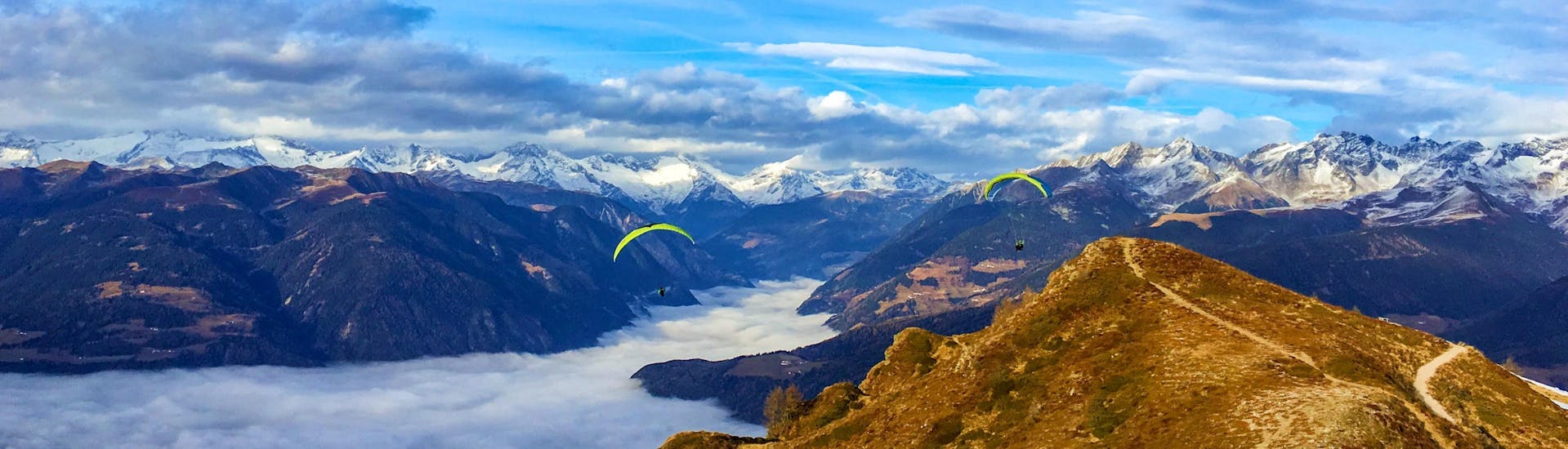 Two paragliders from Tandemflights Kronplatz are gliding over the beautiful mountain scenery around Plan de Corones and the Puster Valley.