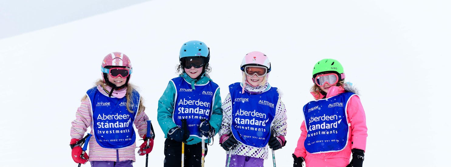 Vier kinderen staan op een rij met helmen en vesten aan tijdens hun skilessen voor kinderen voor alle niveaus bij skischule Altitude in Grindelwald en Wengen.