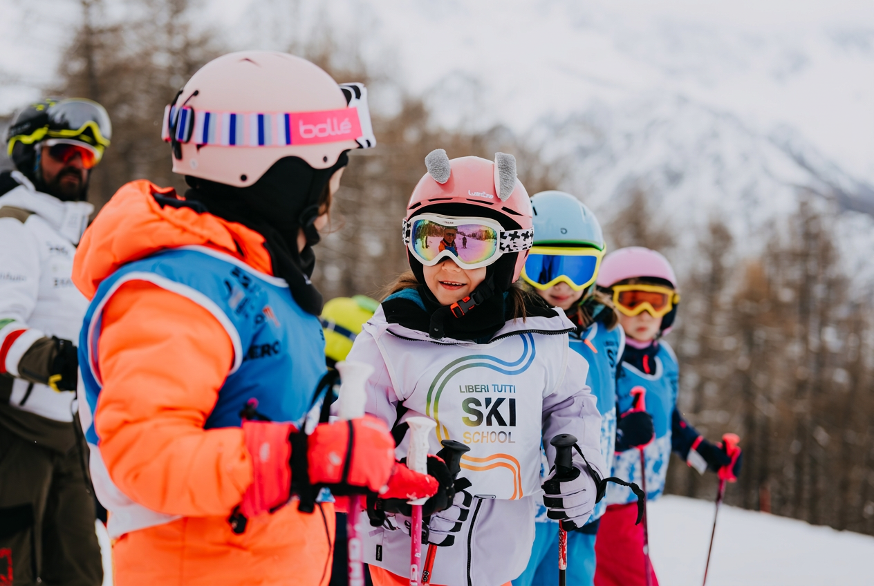 Un gruppo di bambini con caschi e occhiali da sci si trova su un pendio innevato durante una lezione di sci per principianti presso la scuola di sci Liberi Tutti.