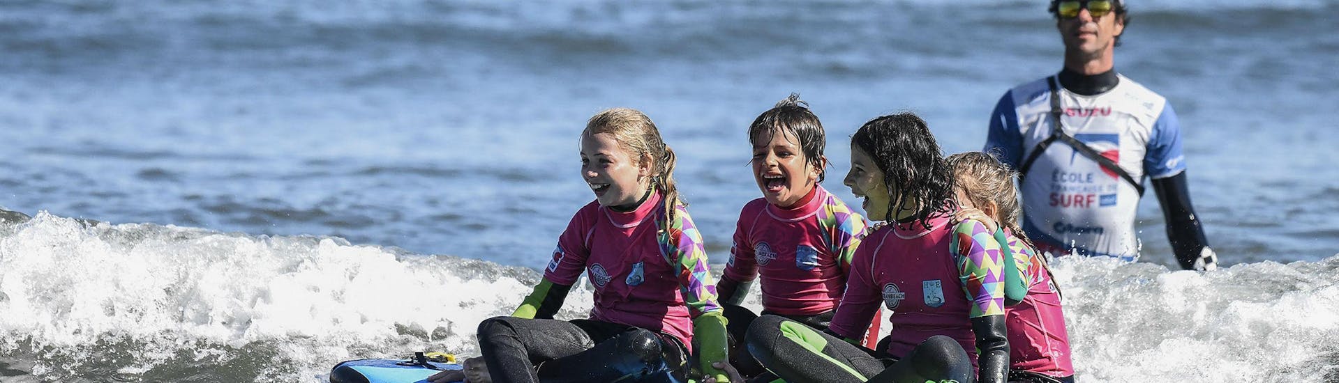 4 niñas se sientan en una tabla de surf en el mar para una lección de surf cerca de su instructor de playa del océano en Hendaya.