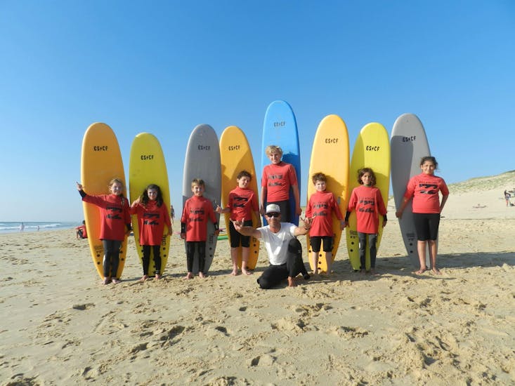 Les enfants sont debout les uns à côté des autres devant leur planche de surf, leur moniteur de surf de l'ESCF Vieux Boucau assis sur le sable, à la fin de leurs cours de surf sur la plage de Sablères.