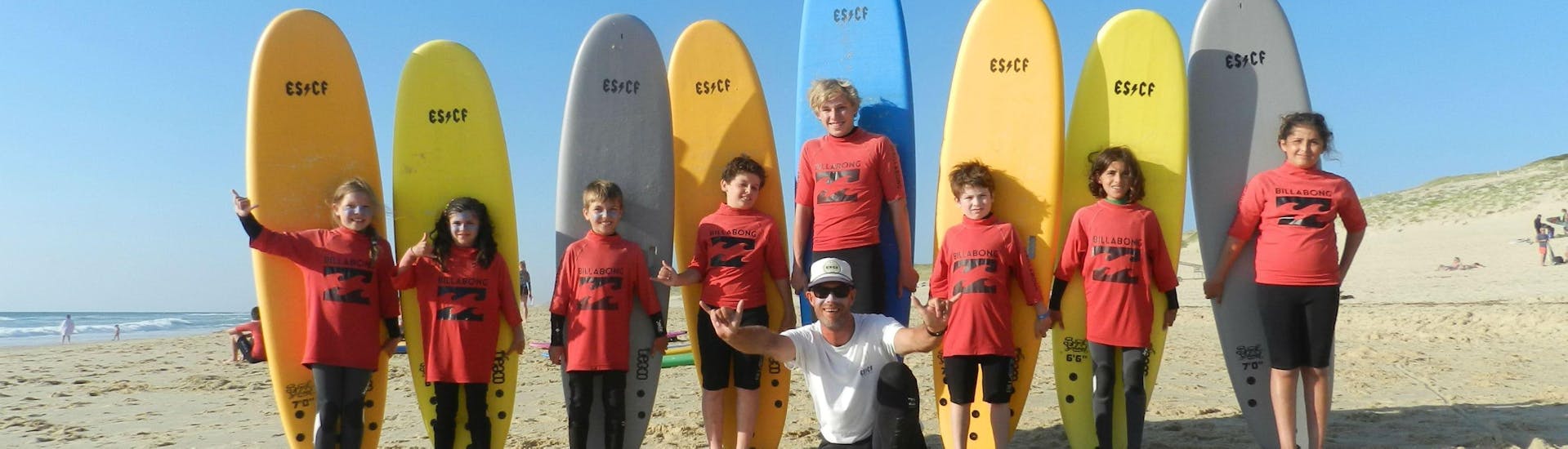 Children are standing next to each other in front of their surboard, their surf instructor from ESCF Vieux Boucau sitting on the sand, at the end of their Surfing Lessons on the Sablères Beach.