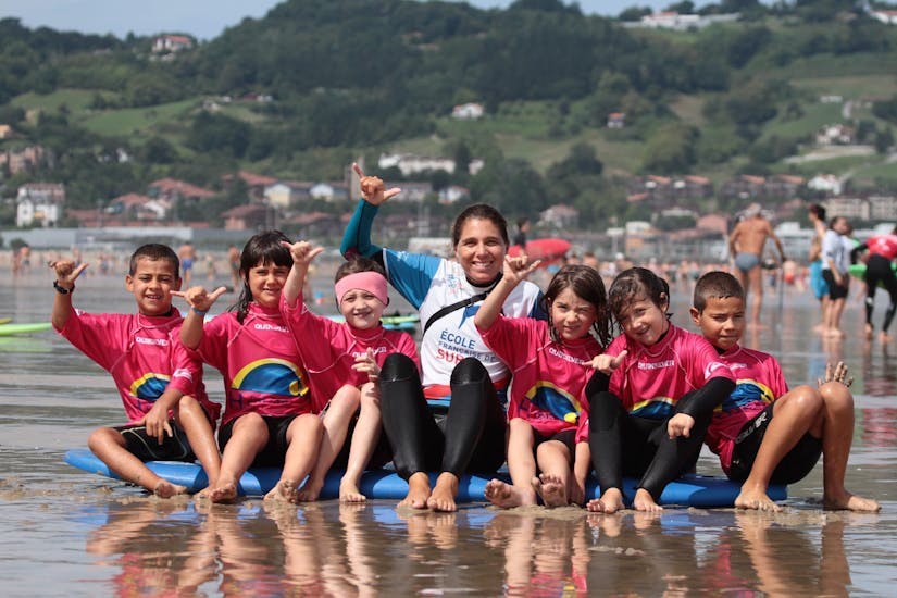 Kids are sitting on the shore on a surf board with their instructor from the surf school École de surf Hendaia during their Surfing Lessons for Kids (6-8 years) on the Hendaye Beach.