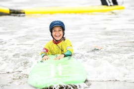 A kid is smiling as they are in the water, holding tight their surfboard, during their Surfing Lessons for Kids (6-8 years) on the Hendaye Beach with École de Surf Hendaia.
