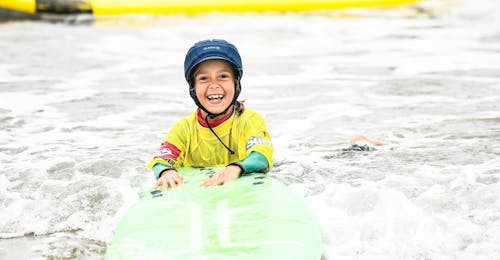 Surflessen voor kinderen (6-8 j.) op het strand van Hendaye met École de Surf Hendaia Een kind lacht terwijl ze in het water liggen, hun surfplank stevig vasthoudend, tijdens hun surflessen voor kinderen (6-8 jaar) op het strand van Hendaye met École de Surf Hendaia.