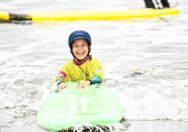 Un enfant sourit alors qu'il est dans l'eau, tenant fermement sa planche de surf, pendant son Cours de surf pour Enfants (6-8 ans) sur la plage d'Hendaye avec l'École de Surf Hendaia.