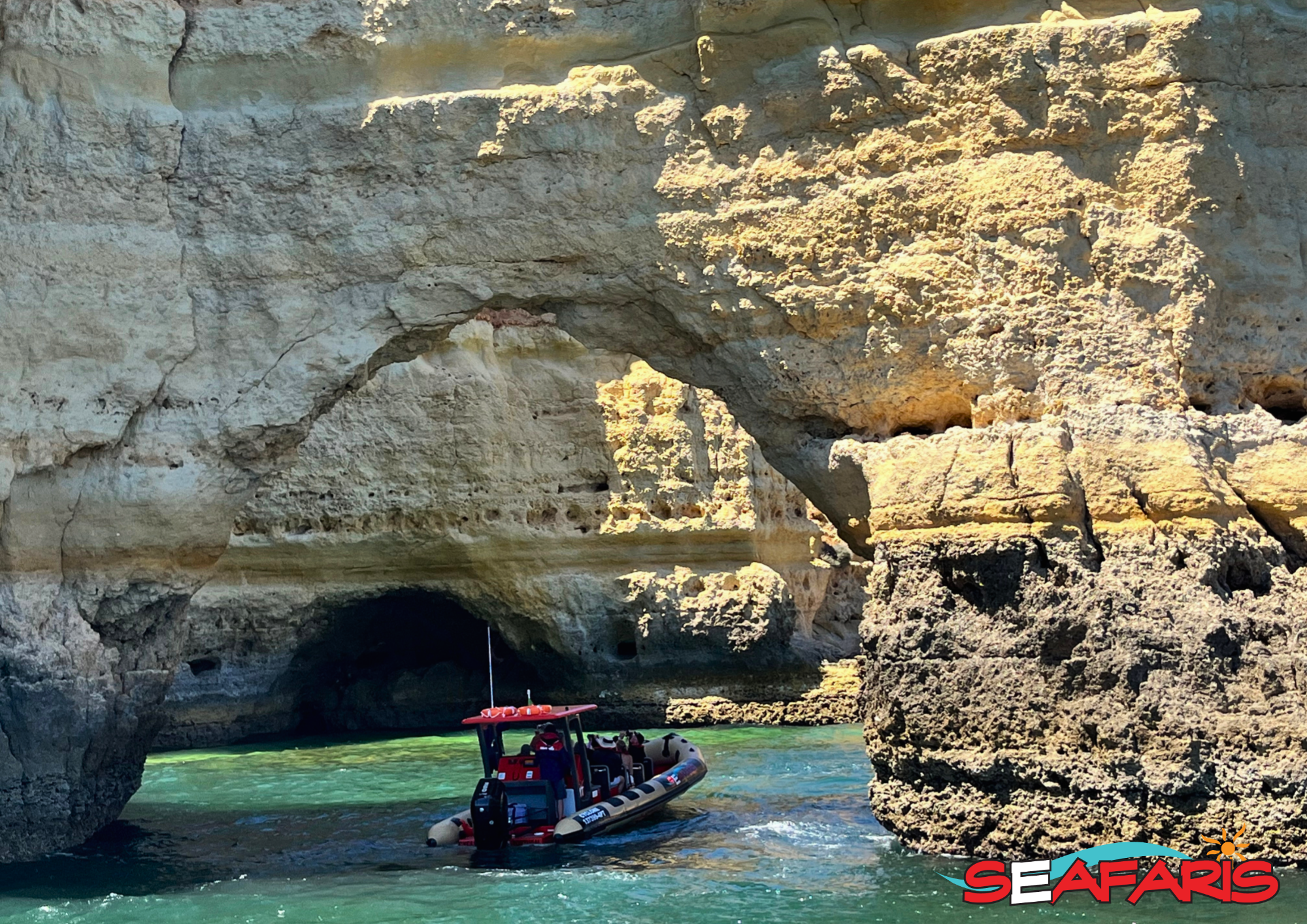 Dolphin Seafaris boat exploring a sea arch in the Algarve. A red tour boat from Dolphin Seafaris Algarve enters a large, sunlit sea cave with turquoise water during a Lagos: Benagil Cave & Sea Caves Boat Tour.