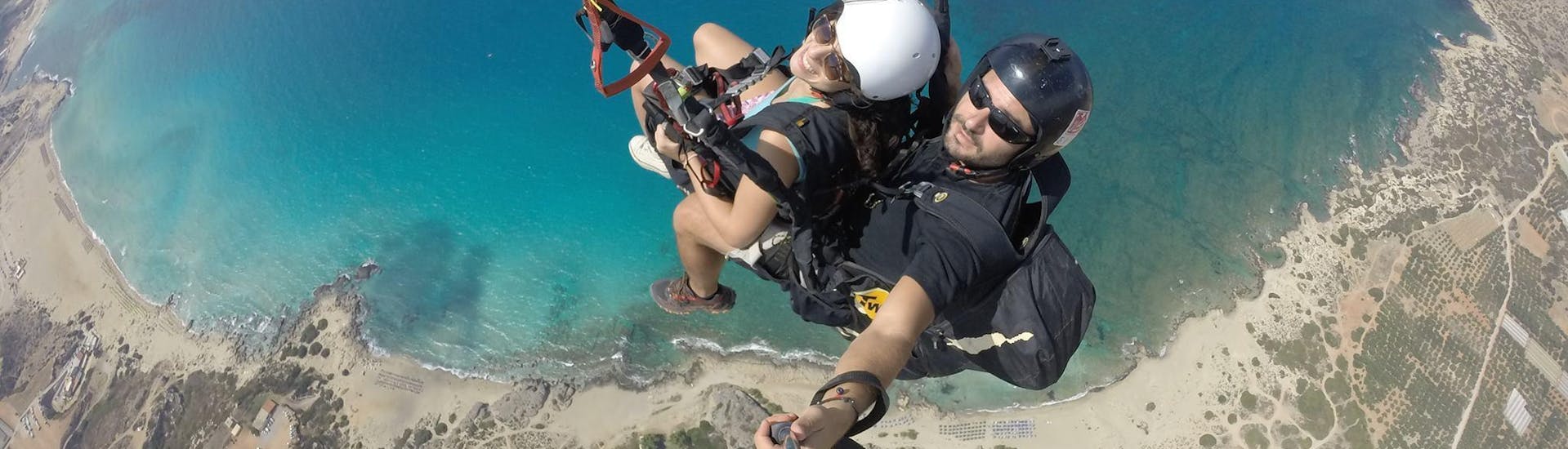 Una mujer sonríe a la cámara mientras vuela en parapente biplaza sobre la playa de Falasarna con un piloto certificado de Cretan Paragliding.