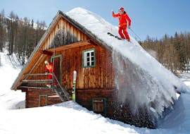 Deux skieurs passent par-dessus le toit d'une cabane pendant leur cours particulier de ski hors-piste pour adultes de tous niveaux avec la skischule Tritscher.