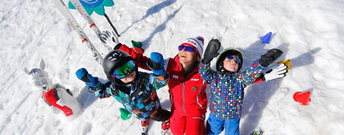 Des enfants sont dans la neige avec les bras levés, à côté de leur moniteur de ski de l'école de ski ESF Alpe d'Huez pendant leur Cours de ski pour Enfants "Chalet des Enfants" (2-5 ans).