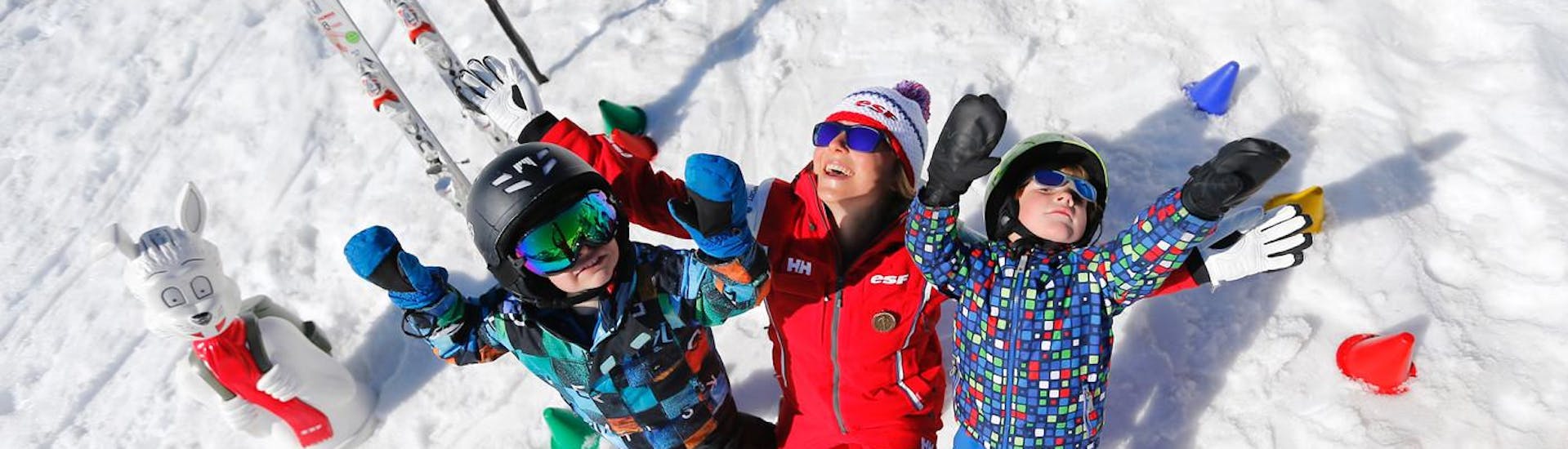 Kinderen staan ​​in de sneeuw met hun armen in de lucht naast hun skileraar van de skischool ESF Alpe d'Huez tijdens hun Kids Ski Lessen "Kinderchalet" (2-5 jaar).